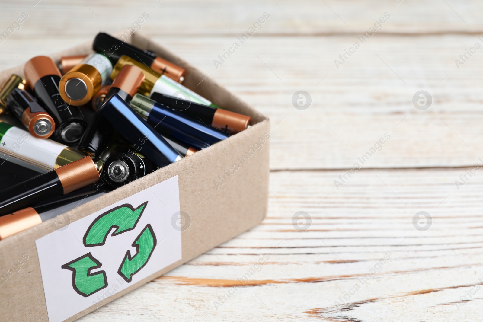 Image of Used batteries in cardboard box with recycling symbol on white wooden table, closeup. Space for text