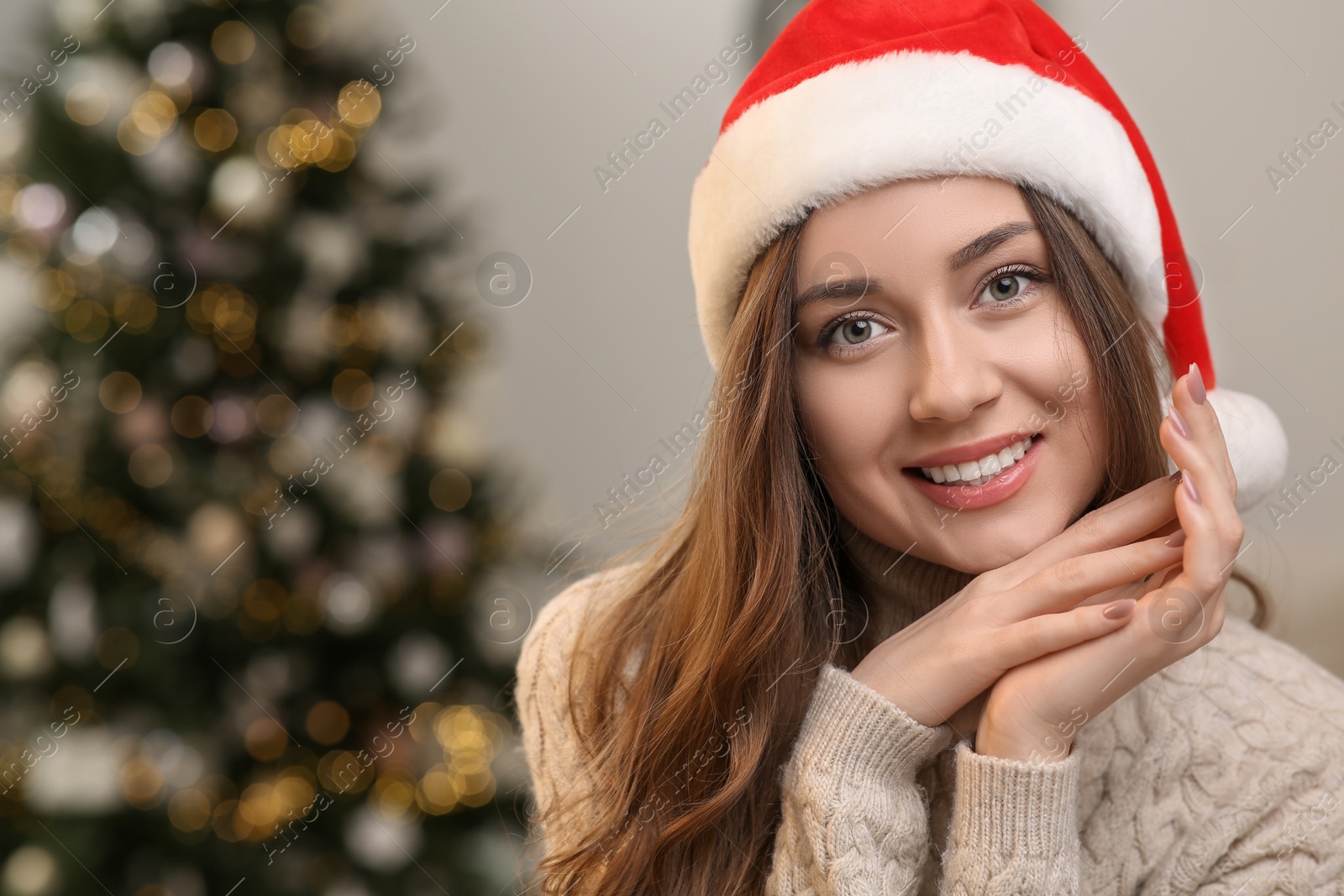 Photo of Beautiful young smiling woman in Santa hat near Christmas tree indoors, space for text