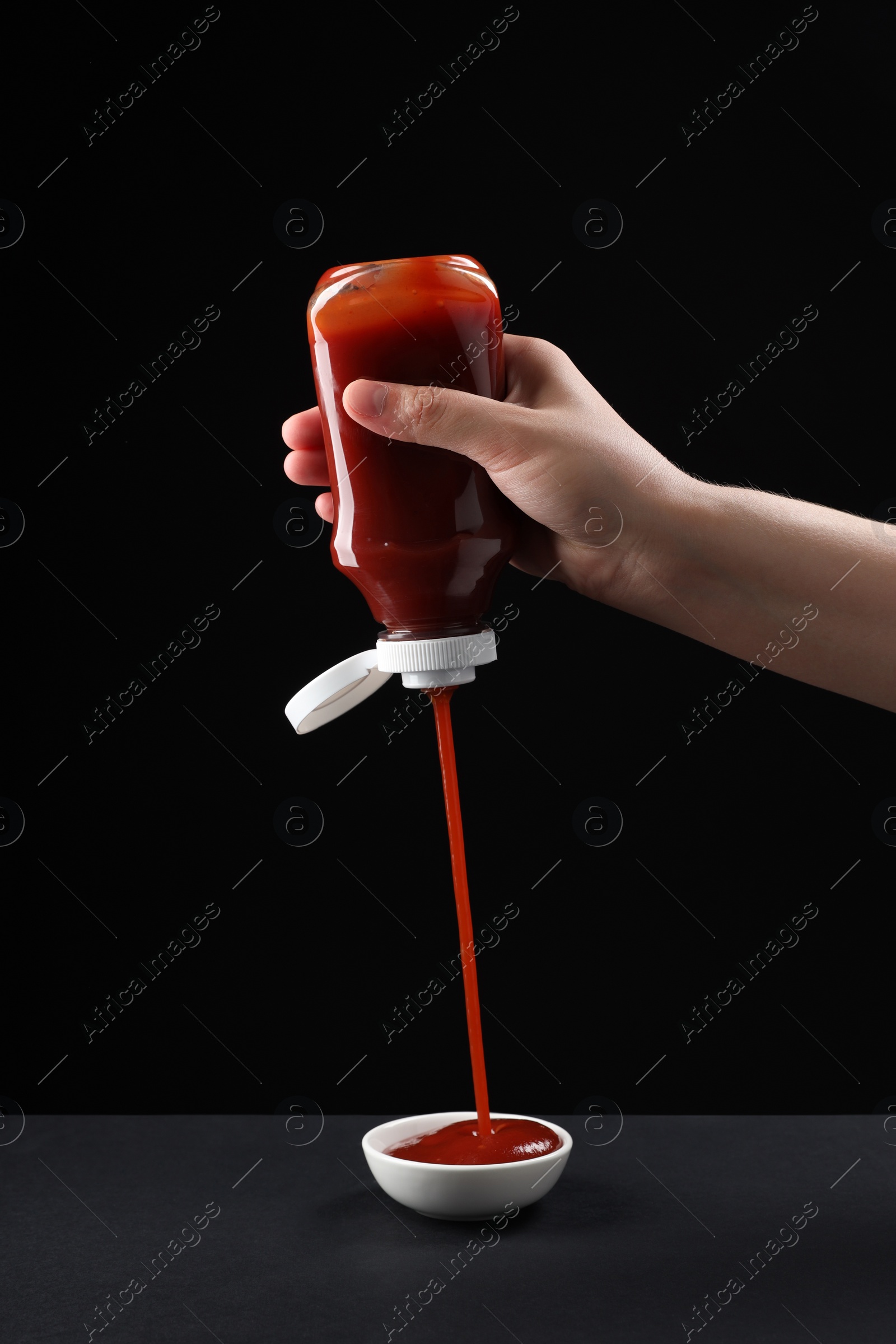 Photo of Woman pouring tasty ketchup from bottle into bowl on table against black background, closeup