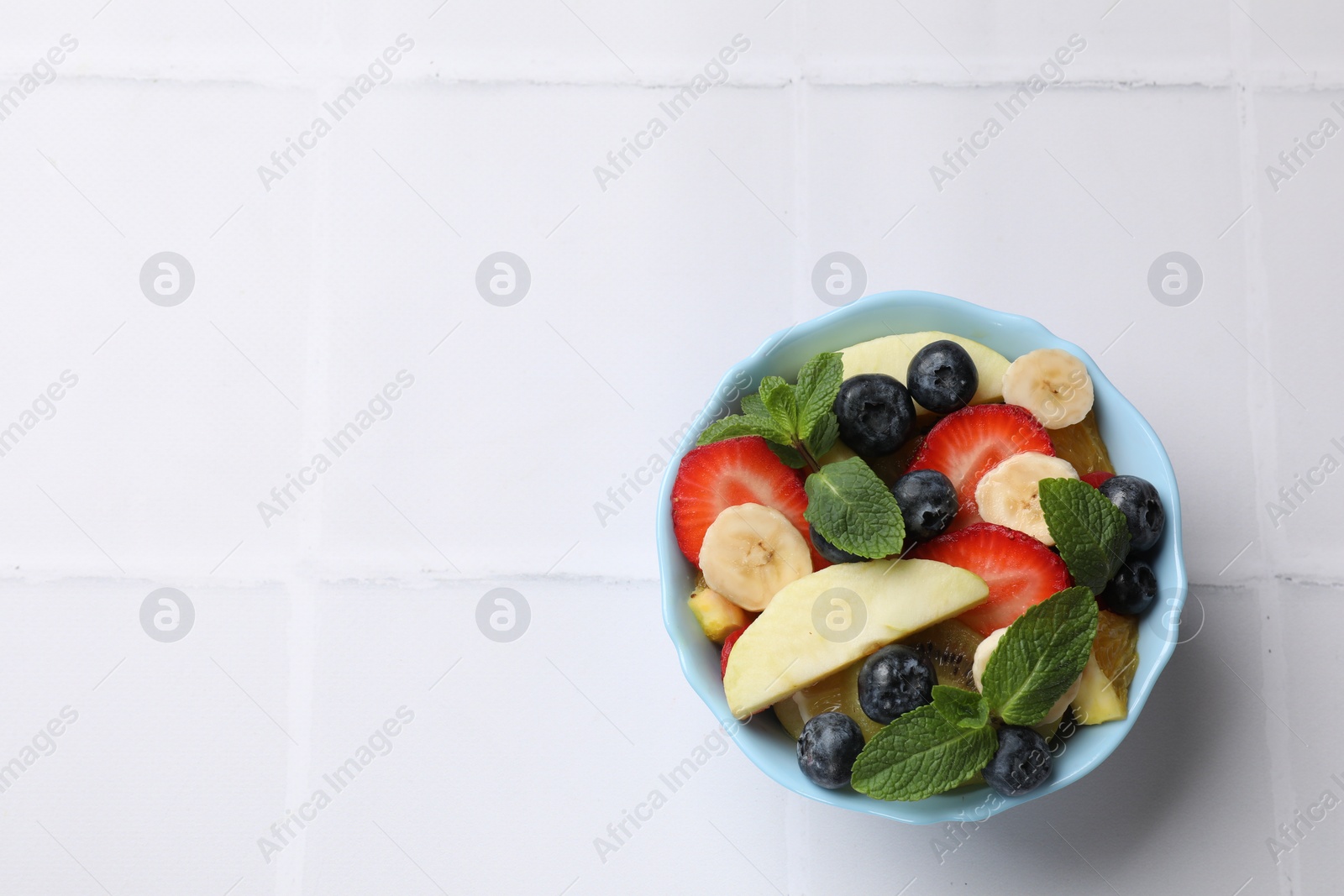 Photo of Tasty fruit salad in bowl on white tiled table, top view. Space for text