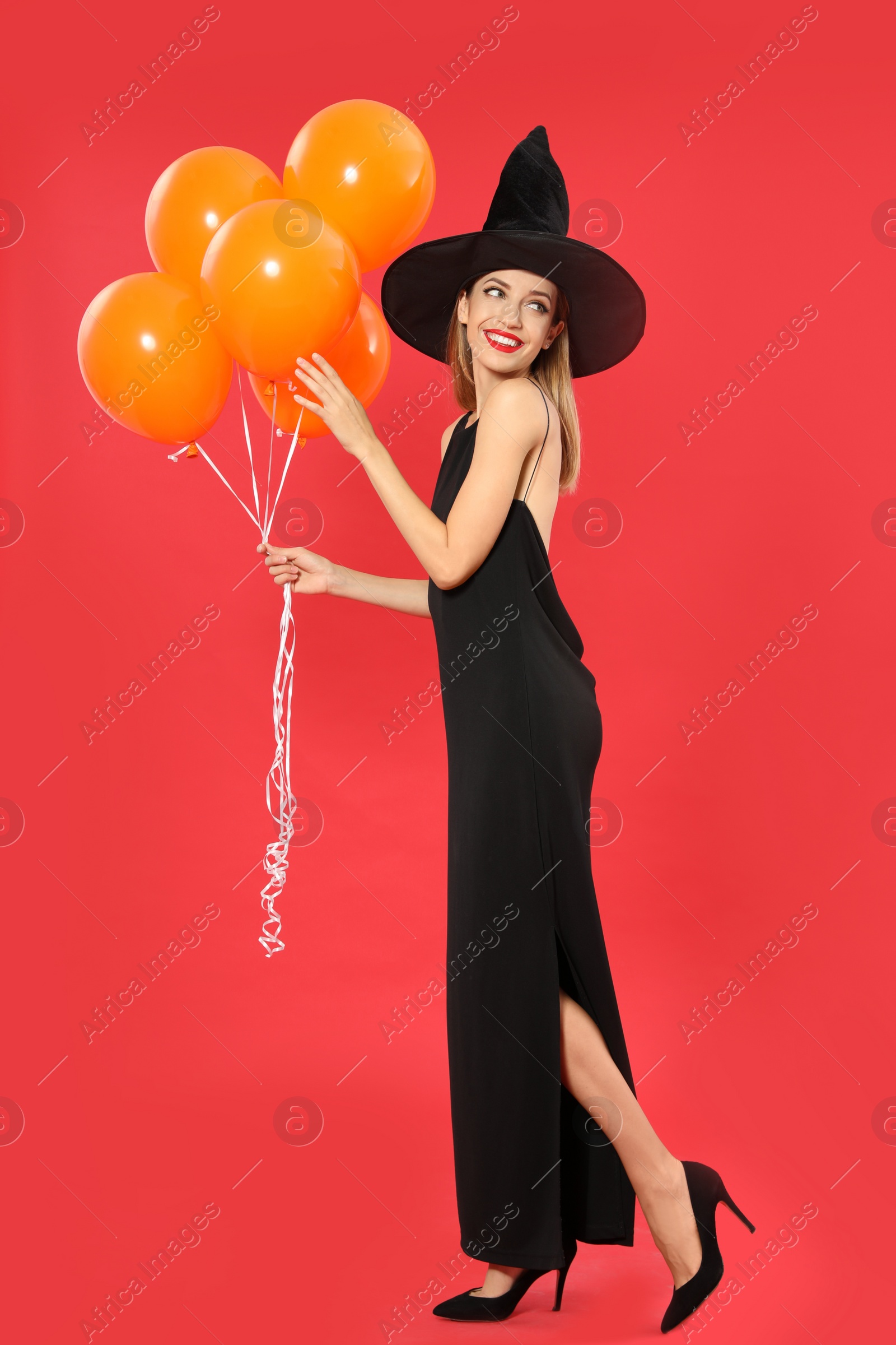 Photo of Beautiful woman wearing witch costume with balloons for Halloween party on red background