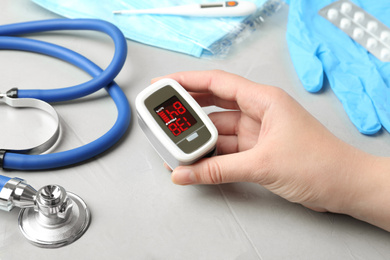 Photo of Woman holding fingertip pulse oximeter near medical items at light grey stone table, closeup