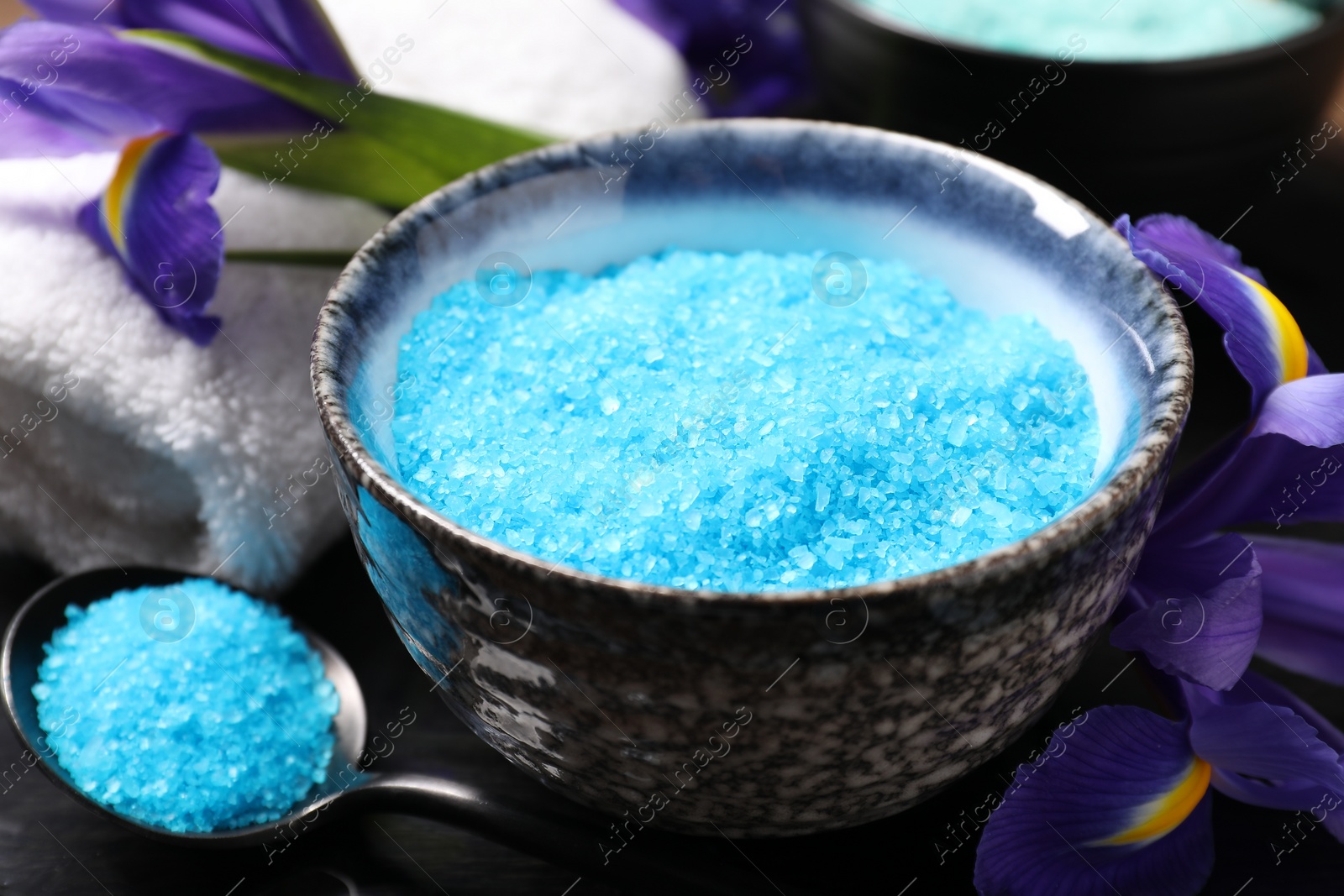 Photo of Light blue sea salt in bowl, flowers, spoon and towel on table, closeup
