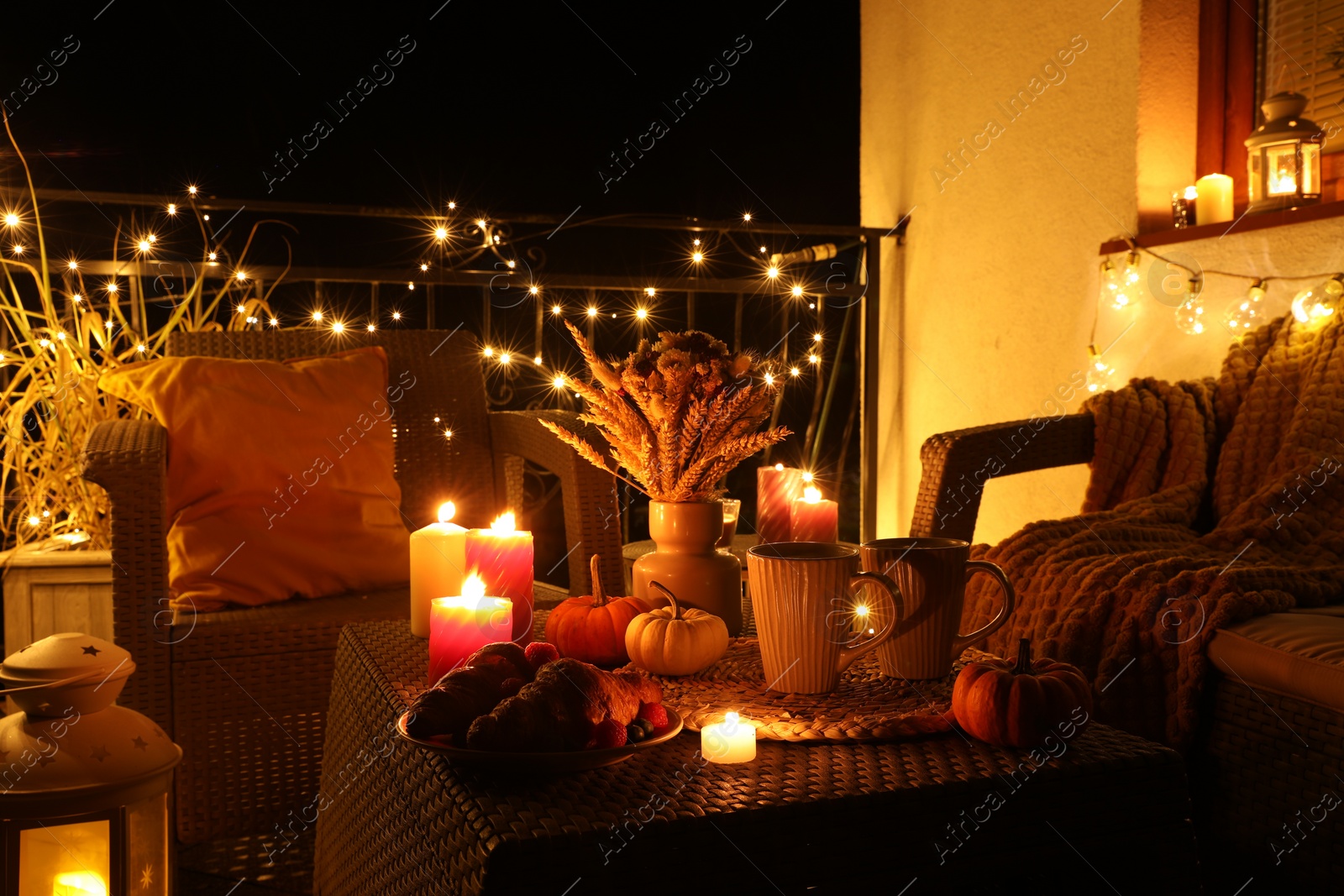 Photo of Rattan furniture, cups, fairy lights, burning candles and other autumn decor on outdoor terrace at night