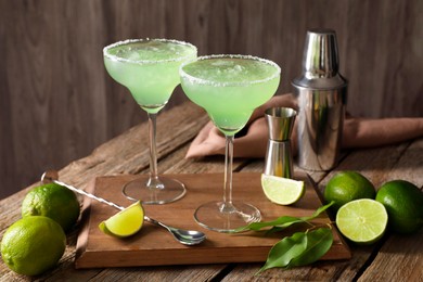 Delicious Margarita cocktail in glasses, limes and bartender equipment on wooden table