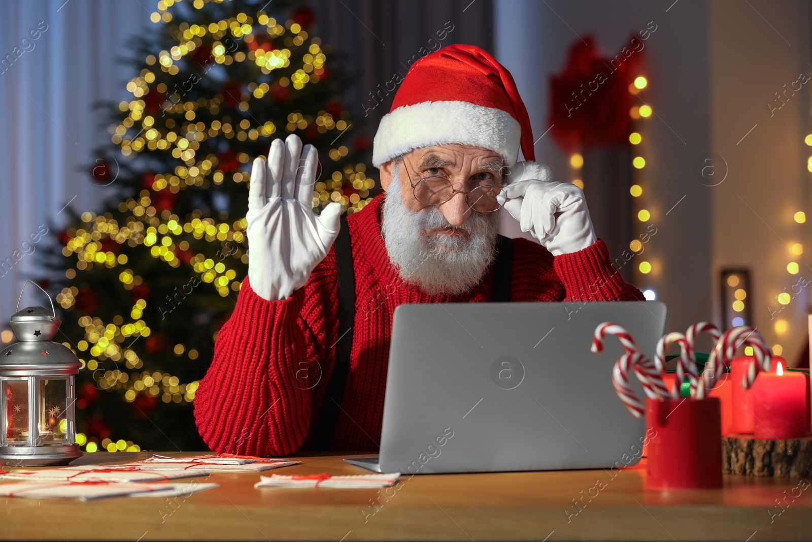 Photo of Santa Claus using laptop and waving hello at his workplace in room with Christmas tree