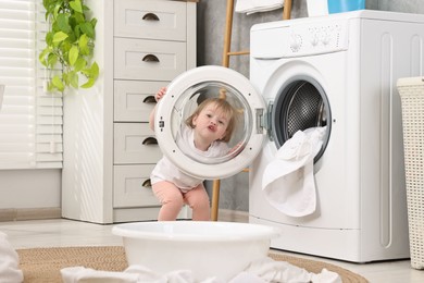 Photo of Little girl having fun near washing machine with baby clothes in bathroom