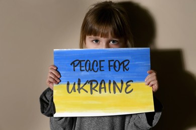 Image of Sad little girl holding paper with phrase Peace for Ukraine near beige wall