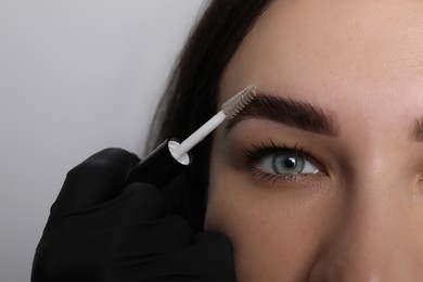 Photo of Beautician fixing woman's eyebrows with gel after tinting on light grey background, closeup