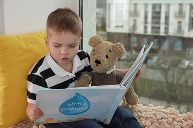 Photo of Cute little boy with toy bear reading book near window at home