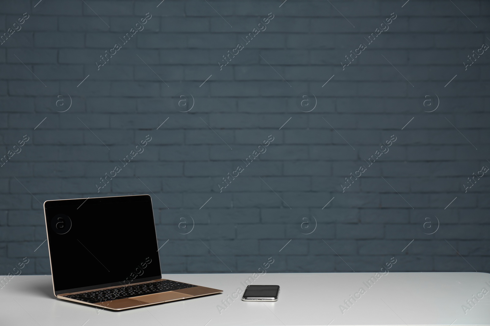 Photo of Modern laptop with blank screen and mobile phone on table against brick wall