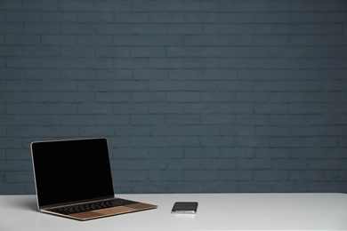 Photo of Modern laptop with blank screen and mobile phone on table against brick wall