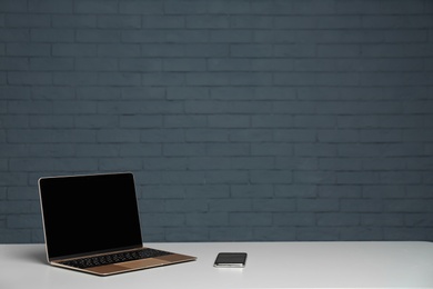 Modern laptop with blank screen and mobile phone on table against brick wall