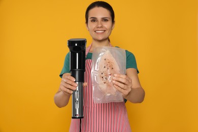 Photo of Beautiful young woman holding sous vide cooker and meat in vacuum pack against orange background, focus on hands