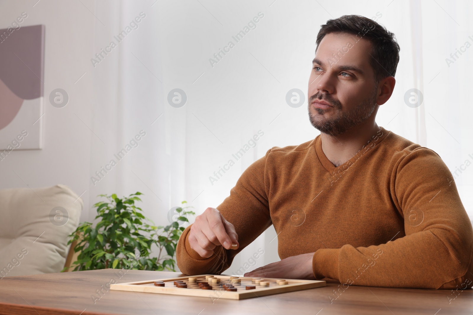 Photo of Playing checkers. Concentrated man thinking about next move at table in room, space for text