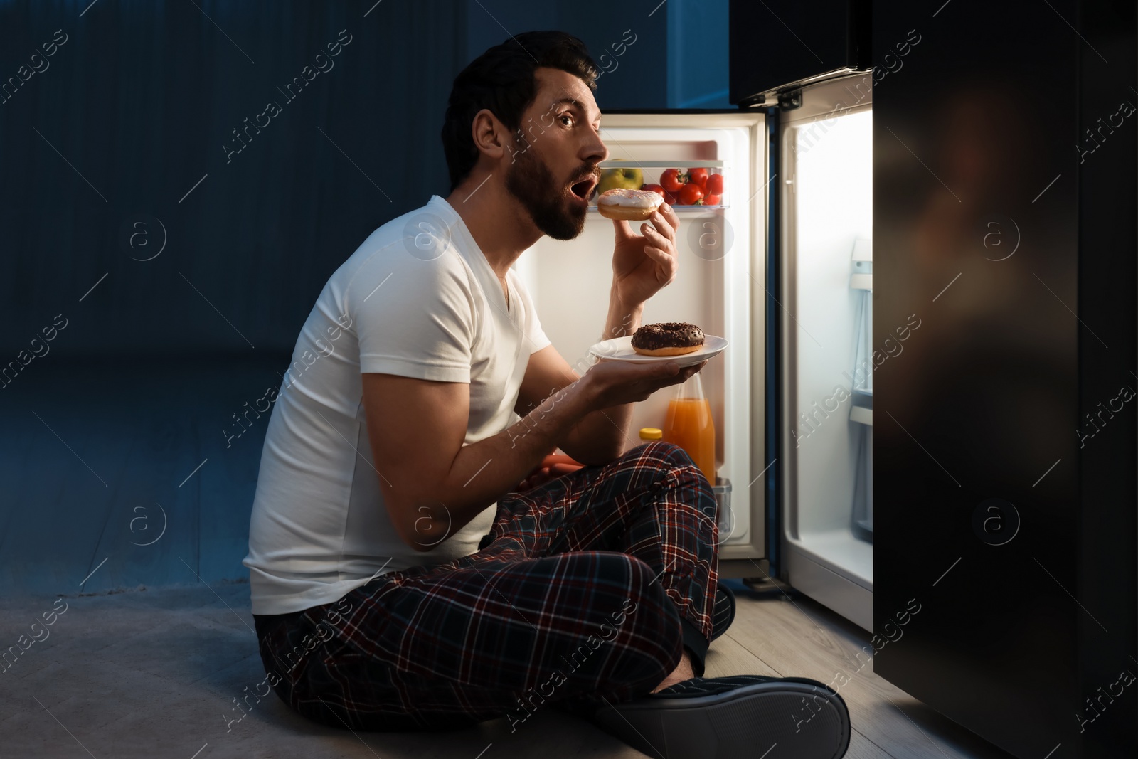 Photo of Man eating donuts near refrigerator in kitchen at night. Bad habit