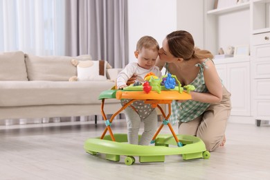 Photo of Cute boy making first steps with baby walker. Mother kissing her little son at home, space for text