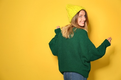 Happy young woman wearing warm sweater and knitted hat on yellow background. Space for text