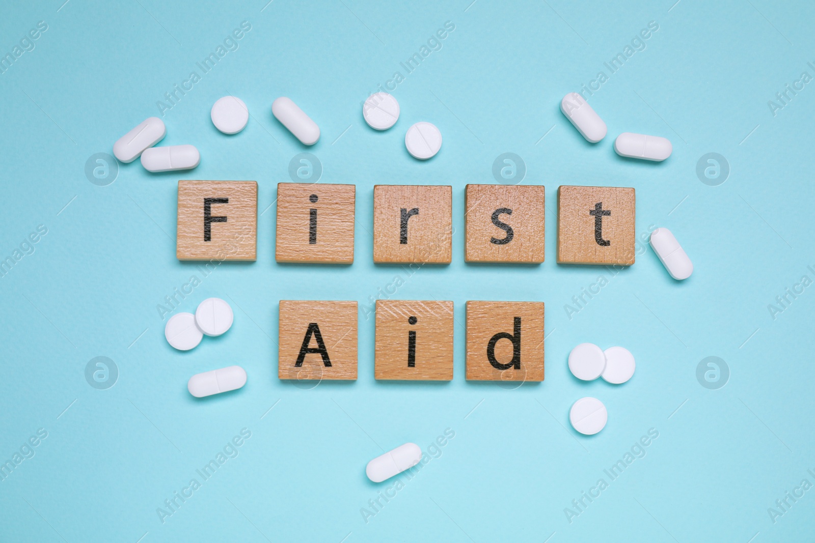 Photo of Words First Aid made of wooden cubes and pills on light blue background, flat lay