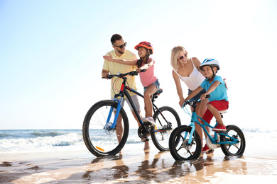 Happy parents teaching children to ride bicycles on sandy beach near sea