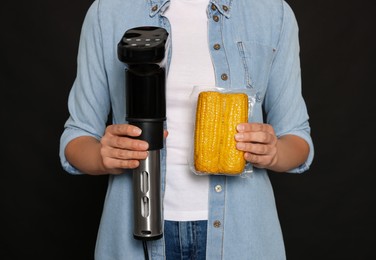 Woman holding sous vide cooker and corn in vacuum pack on black background, closeup