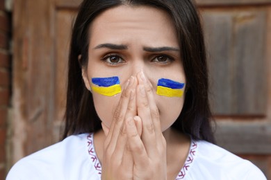 Sad young woman with drawings of Ukrainian flag on face near wooden door, closeup