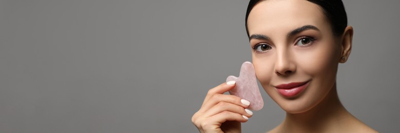 Beautiful young woman doing facial massage with gua sha tool on grey background, space for text. Banner design