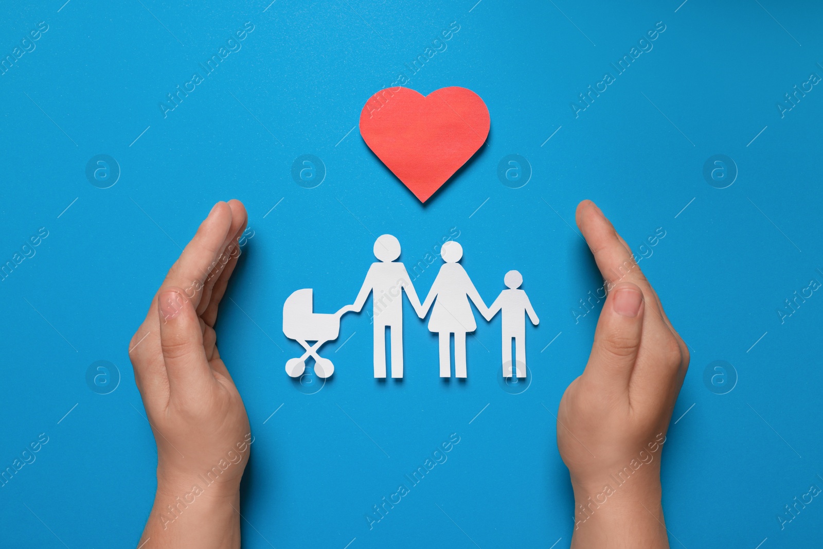 Photo of Man protecting paper family figures and red heart on light blue background, top view. Insurance concept