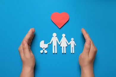 Photo of Man protecting paper family figures and red heart on light blue background, top view. Insurance concept