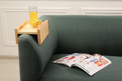 Photo of Glass of juice, chips and magazine on sofa with wooden armrest table in room. Interior element