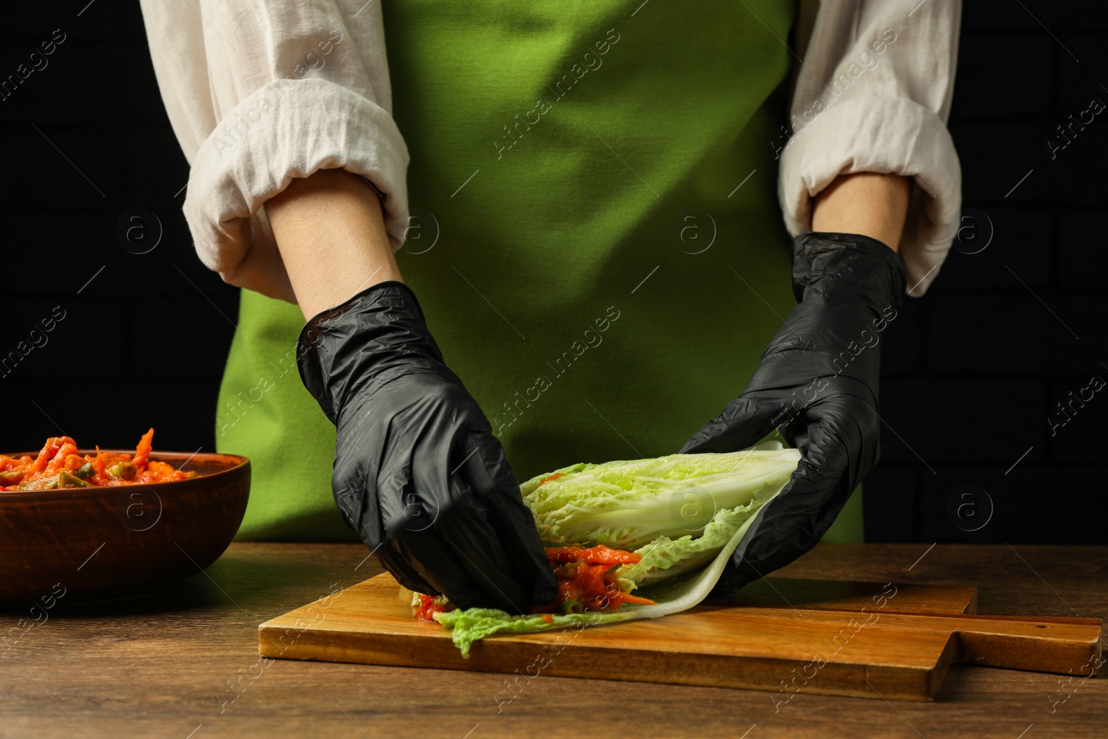 Photo of Woman preparing spicy cabbage kimchi at wooden table against dark background, closeup