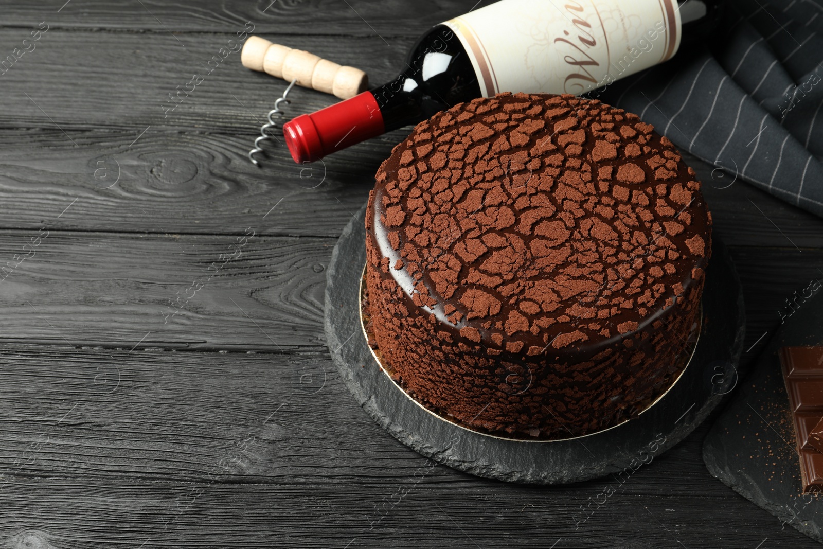 Photo of Delicious truffle cake, bottle of wine, chocolate pieces and corkscrew on black wooden table, space for text