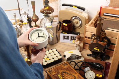 Photo of Woman holding alarm clock near table with different stuff indoors, closeup. Garage sale