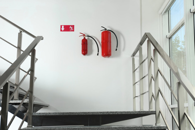 Photo of Fire extinguishers and emergency exit sign on white wall near staircase indoors