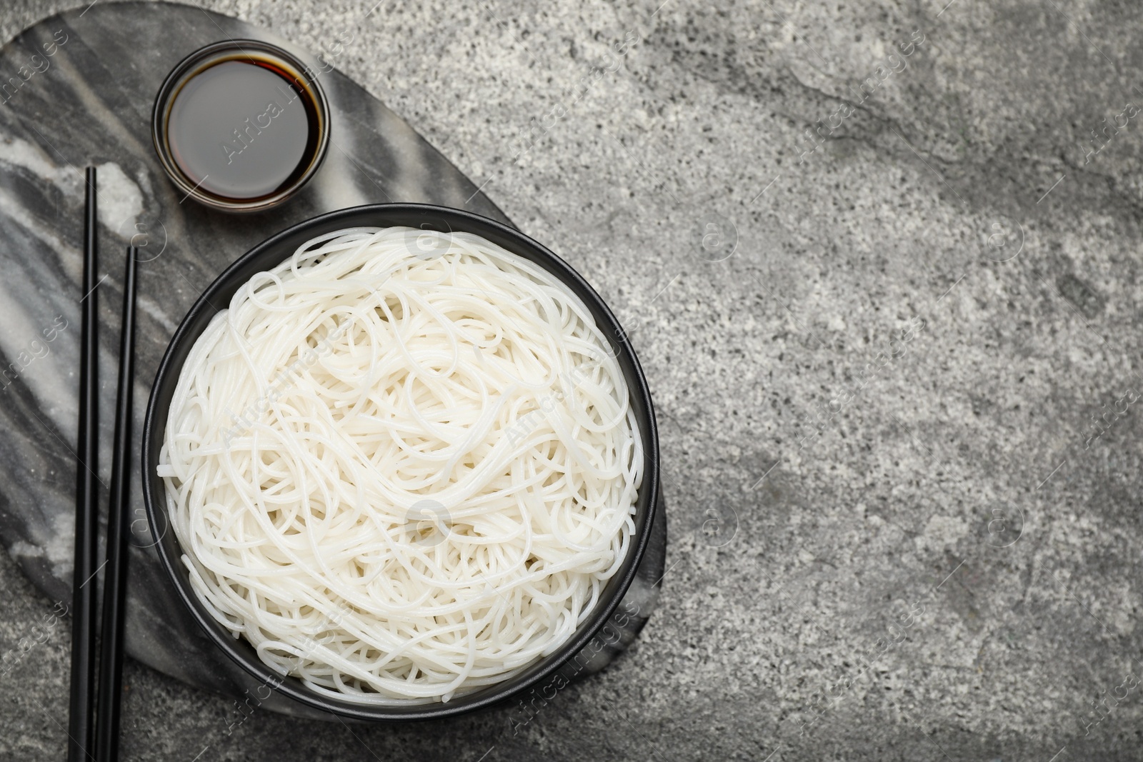 Photo of Bowl with cooked rice noodles, soy sauce and chopsticks on grey table, top view. Space for text