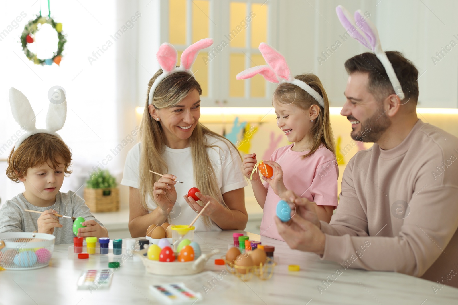 Photo of Easter celebration. Happy family with bunny ears painting eggs at white marble table in kitchen