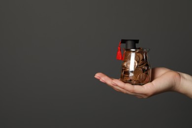 Woman holding glass jar of coins and graduation cap against dark grey background, closeup with space for text. Scholarship concept