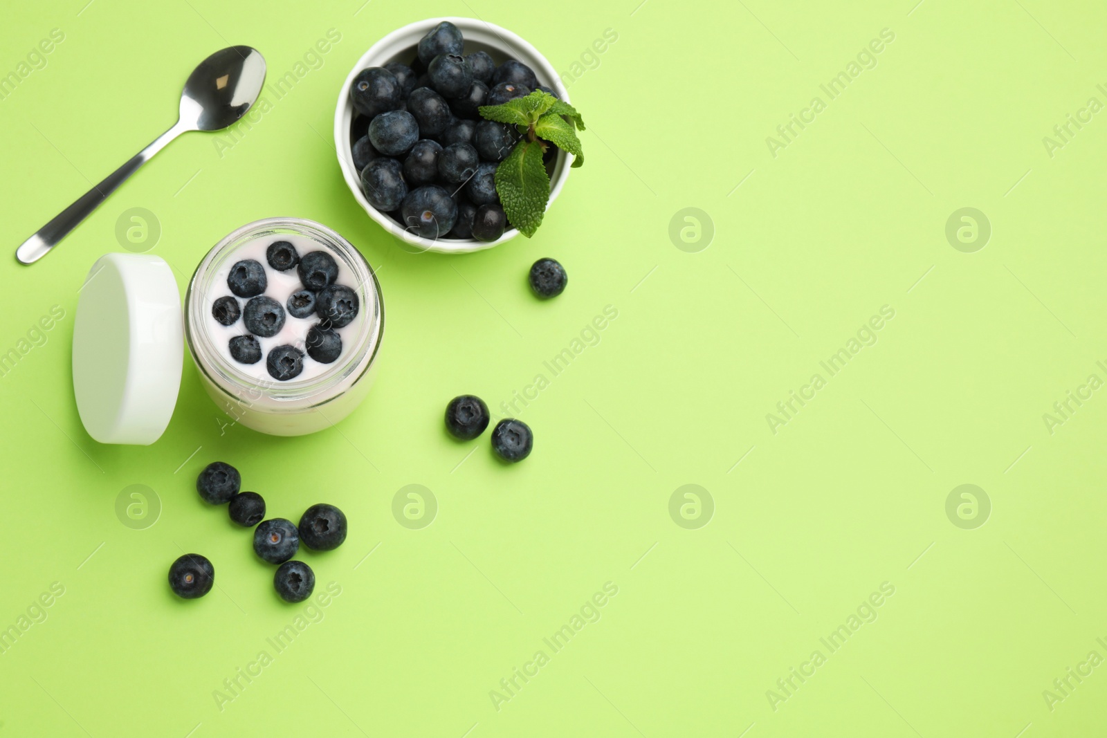 Photo of Jar of fresh yogurt with blueberry and spoon on light green background, flat lay. Space for text