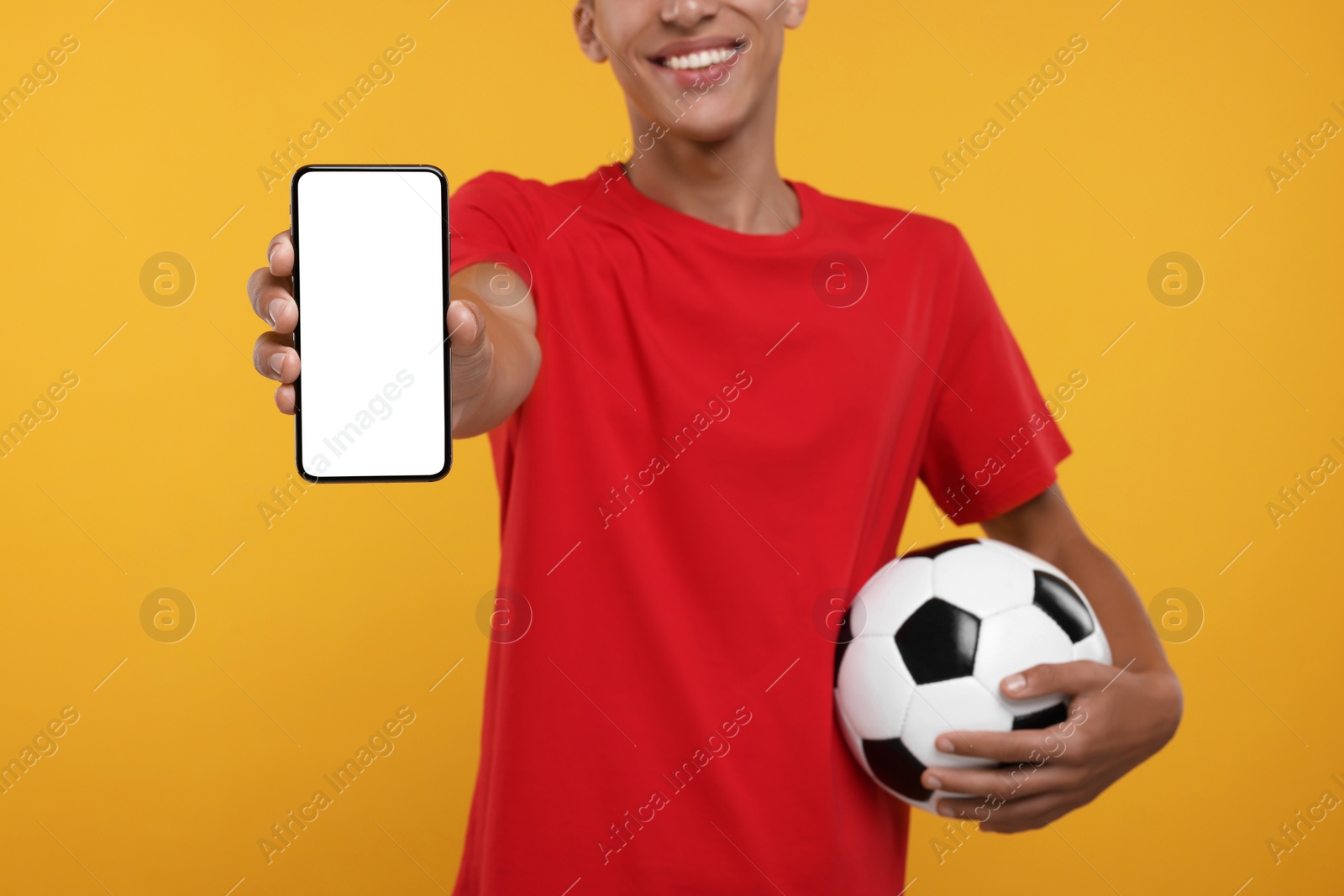 Photo of Happy sports fan with soccer ball showing smartphone on orange background, closeup