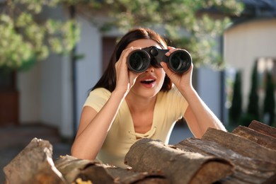 Photo of Concept of private life. Curious young woman with binoculars spying on neighbours over firewood outdoors
