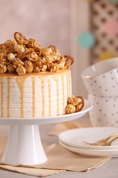 Photo of Caramel drip cake decorated with popcorn and pretzels near tableware on light grey table