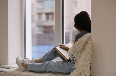 Cute little girl reading book near window at home