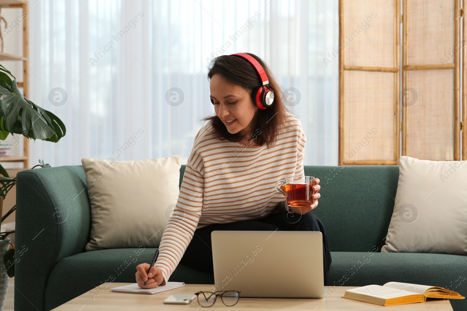 Photo of Woman with modern laptop and headphones drinking tea while learning at table indoors