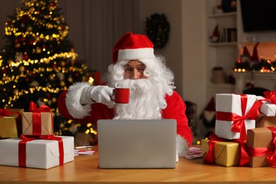 Photo of Santa Claus holding cup of drink using laptop at table with Christmas gifts in room