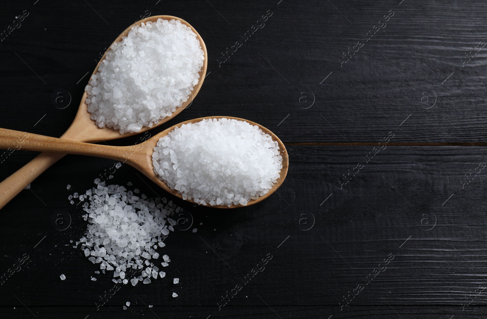 Photo of Organic salt in spoons on black wooden table, top view. Space for text