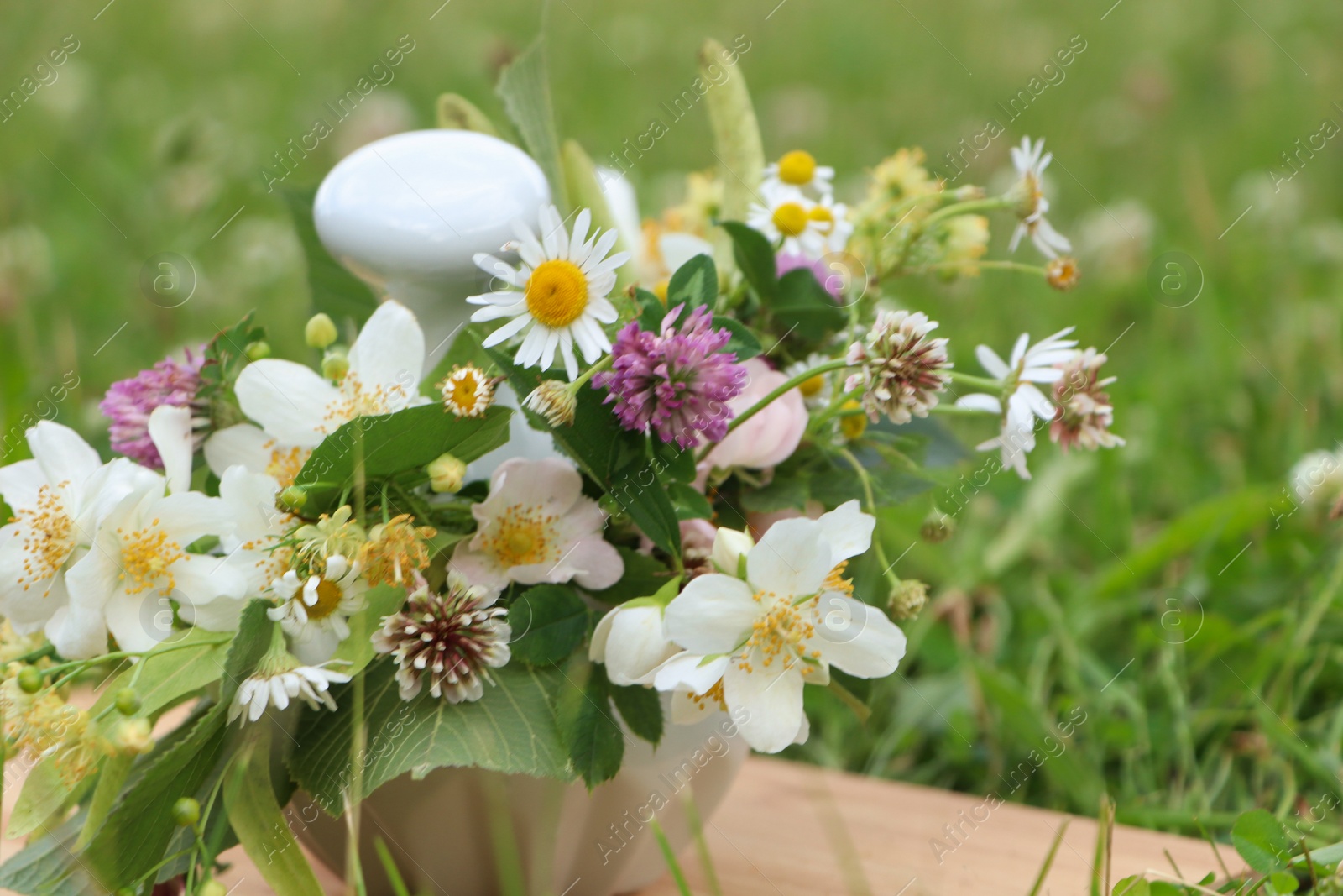 Photo of Ceramic mortar with pestle, different wildflowers and herbs on wooden board in meadow, closeup