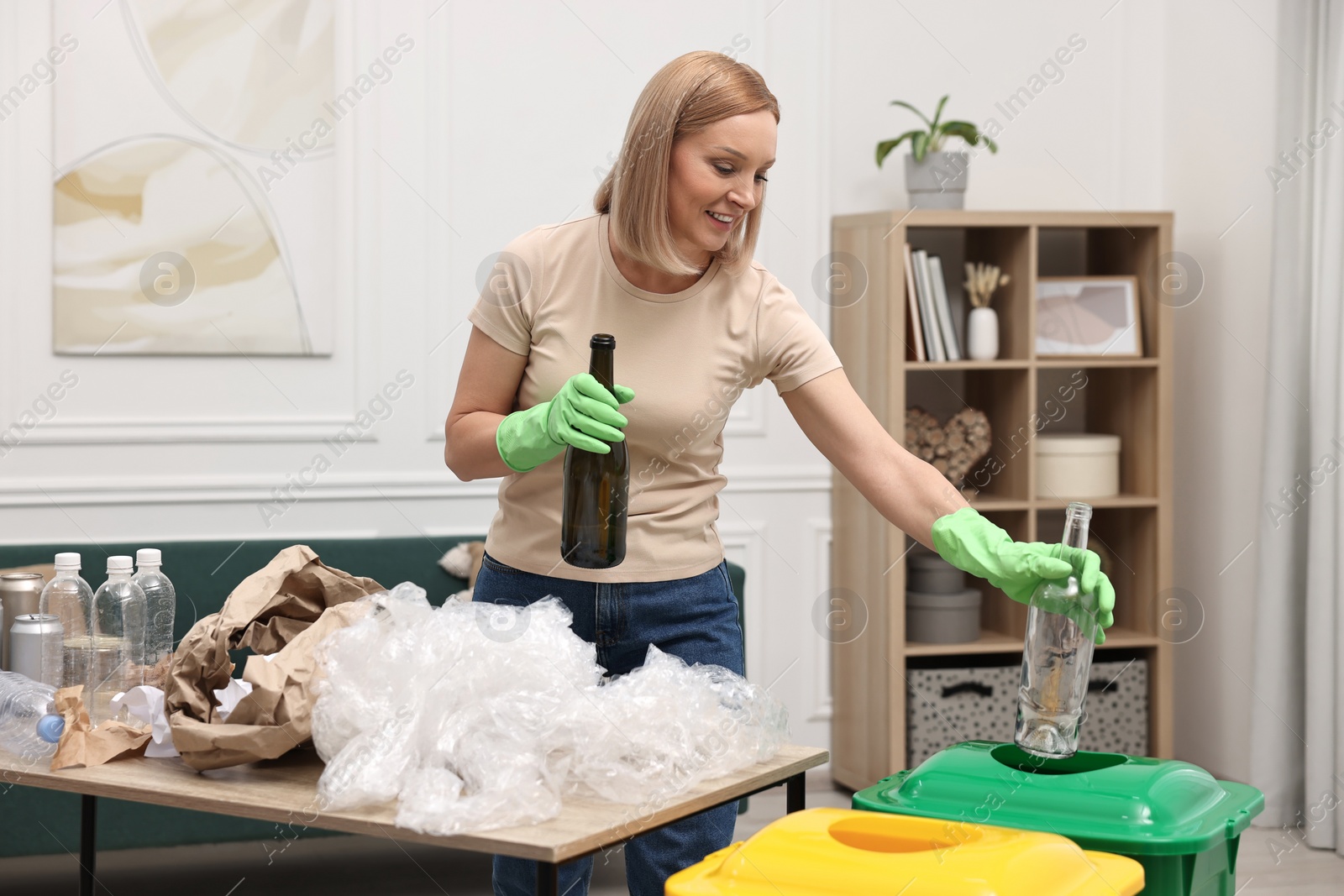 Photo of Garbage sorting. Smiling woman throwing glass bottle into trash bin in room