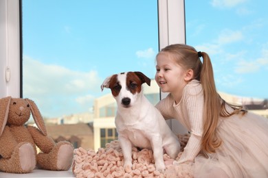 Photo of Cute little girl with her dog sitting on window sill indoors, space for text. Childhood pet