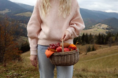 Photo of Woman holding wicker picnic basket with fruits and autumn leaves in mountains, closeup