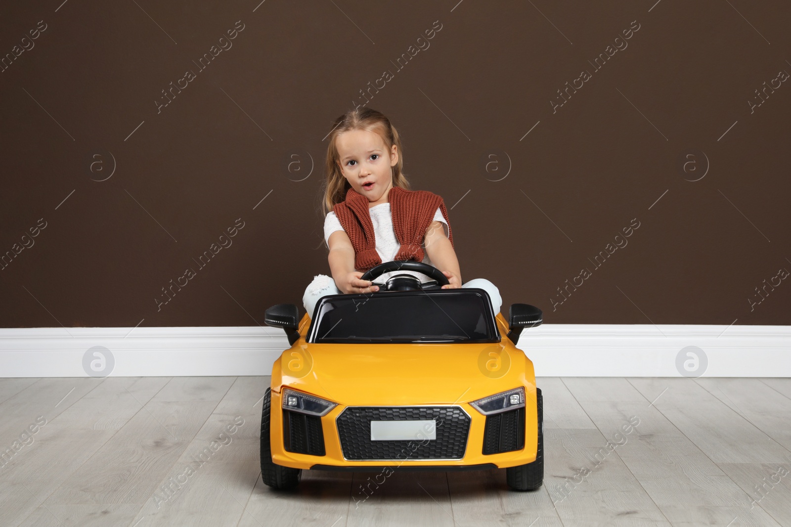 Photo of Cute little girl driving children's electric toy car near brown wall indoors